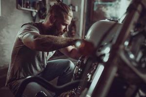 hombre guapo con barba reparando su motocicleta en el garaje. un hombre vestido con jeans y una camiseta foto