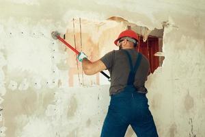 builder with a hammer in his hands breaks the cement wall The builder is dressed in a protective suit and helmet photo