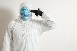 a man in a protective suit hung with medical masks posing against a wall background showing various gestures with his fingers the scientist gestures a pistol to his temple