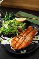 un trozo de salmón al horno, pimienta, limón y sal sobre un plato negro con hojas de lechuga sobre un fondo de piedra foto