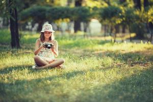 la niña pequeña es interpretada por una cámara fotográfica sentada en el césped en el parque. haciendo selfie y fotografiando el mundo alrededor foto