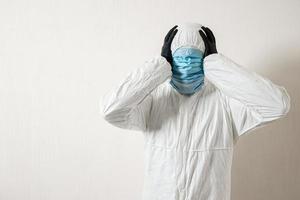 a man in a protective suit hung with medical masks posing against a wall background showing various gestures with his fingers photo