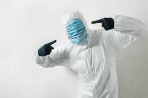 man in a protective suit posing against wall background showing various gestures with his fingers. scientist gestures with his hands to indicate a set of medical masks on his face photo