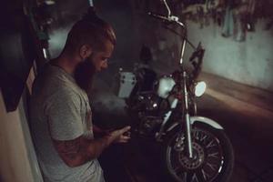 un apuesto hombre brutal con barba está parado en su garaje contra el fondo de una motocicleta y mirando hacia otro lado foto