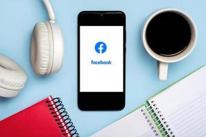 Facebook logo on white screen of smartphone with notebooks,headphone and cup of coffee