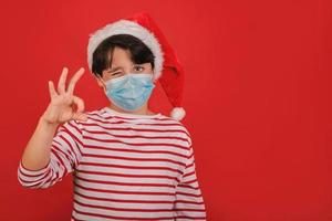 Merry Christmas,kid with medical mask wearing Santa Claus hat showing ok gesture photo