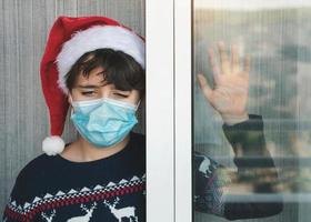 sad kid with medical mask wearing Santa Claus hat wait for santa claus next to a window in Christmas