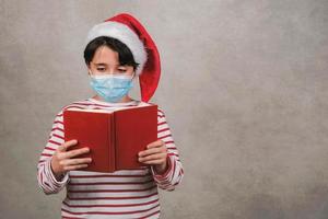 feliz navidad, niño con máscara médica con sombrero de santa claus de navidad leyendo un libro foto