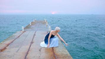 Caucasian beautiful stylish blonde woman in blue and white sitting near the sea at the sunset - protect water, save nature photo