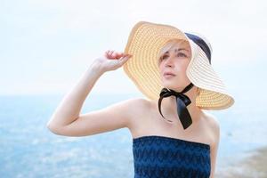 caucásica hermosa y elegante mujer rubia triste vestida de azul y con sombrero de paja parada cerca del mar foto