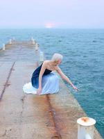 Caucasian beautiful stylish blonde woman in blue and white sitting near the sea at the sunset - protect water, save nature photo