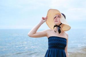 Caucasian beautiful stylish blonde sad woman in blue dress and in straw hat standing near the sea photo