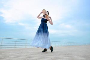 Caucasian beautiful stylish blonde sad woman in blue dress and in straw hat standing by the sea photo