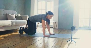 Pretty woman blogger in tracksuit stands in plank pose shooting video with phone in living room during quarantine slow motion