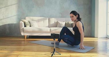 brunette vrouw trainer gaat op mat liggen om nieuwe video voor blog op te nemen in ruime woonkamer verlicht door zonlicht in quarantaine slow motion