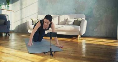 Strong female fitness trainer does push ups posing for camera on smartphone in sunny living room during quarantine slow motion video