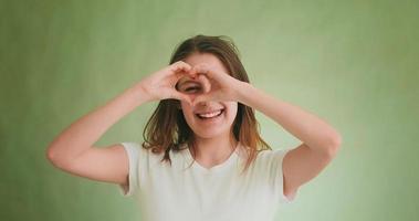 Cheerful young woman in white t-shirt looks through heart made with fingers posing for camera on green closeup slow motion video