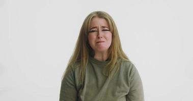 Mujer rubia emocional con sudadera verde llora mirando a la cámara junto a la pared del estudio blanco en la audición a cámara lenta video