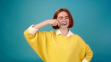 Young woman in yellow sweater brushes teeth with toothbrush and smiles cheerfully posing for camera on blue slow motion close view