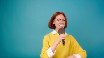 Young woman model with red hair in yellow sweater sings into black hairbrush as microphone in blue studio slow motion closeup