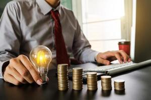 business hand holding lightbulb with using laptop computer and money stack in office. idea saving energy and accounting finance concept  in morning light photo