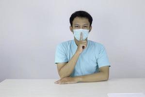 Young asian Man withwearing a surgical mask brings his hand to his nose as a sign of silence, isolated on white background photo