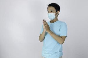 un joven asiático con máscara da las manos de saludo namaste. hombre indonesio sobre fondo gris. atención médica y concepto médico para evitar el virus de la corona. foto