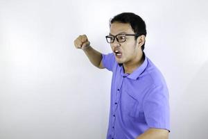 El joven asiático que usa camisa azul es una cara graciosa y enojada con gritos y señalando con el dedo a la cámara aislada sobre el fondo blanco. foto