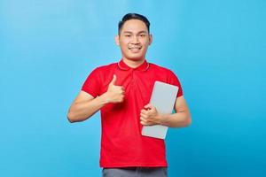 Portrait of smiling Asian young man in red shirt holding laptop and pointing thumbs up while looking at camera isolated on blue background photo