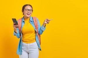 Portrait of  cheerful young Asian woman student in casual clothes with backpack holding mobile phone and pointing at copy space with fingers isolated on yellow background photo