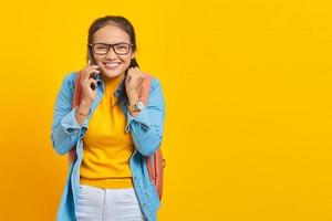 alegre joven estudiante asiática vestida de forma informal con mochila sosteniendo un teléfono móvil, mostrando un gesto de suerte de celebración aislado en un fondo amarillo. educación en concepto de universidad universitaria foto