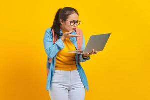 Cheerful young Asian woman student in casual clothes with backpack using laptop and sending air kiss to boyfriend during video call on yellow background. Education in university college concept photo