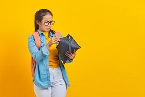 Portrait of young Asian woman student in denim clothes, glasses with backpack, surprised expression while opening wallet isolated on yellow background photo