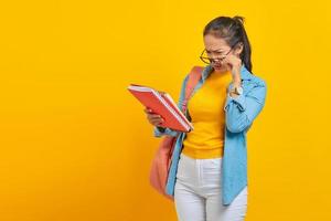 Shocked Beautiful young Asian woman student in denim clothes with backpack, reading notebook and looking at task list isolated on yellow background. Education in high school university college concept photo