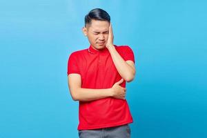 retrato de un apuesto joven asiático con camisa roja que parece mareado y sufre de dolor de cabeza aislado de fondo azul foto