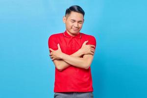 retrato de un alegre joven asiático con camisa roja abrazándose y sintiéndose frío aislado en un fondo azul foto