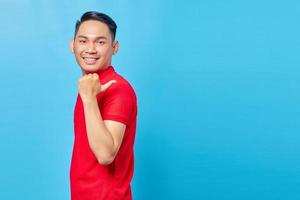 retrato de un joven asiático sonriente con camisa roja de pie señalando con el dedo en el espacio de copia aislado de fondo azul foto