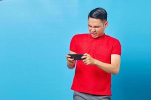 Portrait of disappointed young asian man playing video game on mobile phone isolated on blue background photo