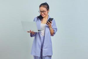 Portrait of smiling young Asian woman looking at laptop and holding mobile phone isolated on white background photo