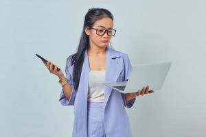 Portrait of young Asian woman standing using laptop and holding mobile phone with pensive expression isolated on white background photo