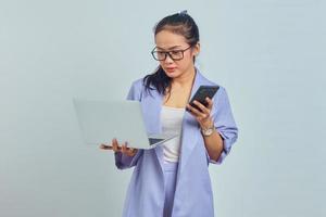 Portrait of pensive young Asian woman looking at laptop and holding mobile phone isolated on white background