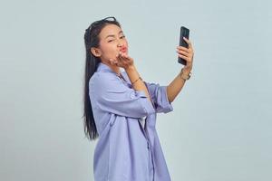 Portrait of charming young Asian woman holding hands sending air kiss to boyfriend while making video call on white background photo