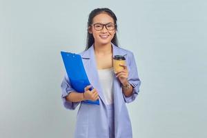 Portrait of cheerful young Asian business woman holding clipboard and drinking takeaway coffee isolated on purple background photo