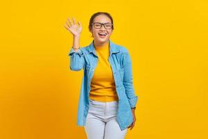 Portrait of cheerful young Asian woman student in denim clothes waving hand at camera isolated on yellow background. People sincere emotions lifestyle concept photo