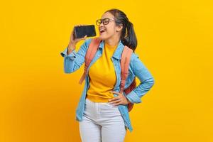 retrato de una joven estudiante asiática sonriente vestida de forma informal con mochila disfrutando cantando con un smartphone aislado de fondo amarillo. educación en concepto de universidad universitaria foto