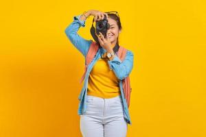 retrato de una alegre joven asiática con mochila tomando fotos en una cámara profesional aislada de fondo amarillo