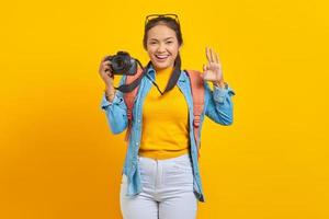 Portrait of smiling young Asian woman with backpack holding professional camera and showing okay gesture isolated on yellow background photo