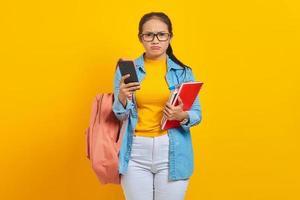 Portrait of pensive young Asian woman student in denim clothes with backpack holding mobile phone and books isolated on yellow background photo