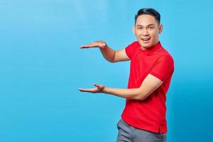 Handsome Asian young man in red shirt smiling showing product with palm, showing copy space for certain discount isolated over blue background photo