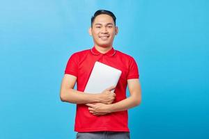 Portrait of smiling Asian handsome young man in red shirt hugging laptop isolated on blue background photo